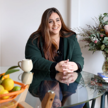 Smiling woman with long brown hair, wearing a dark green jacket, seated at a glass table with a mug, fruit bowl, and flowers nearby.